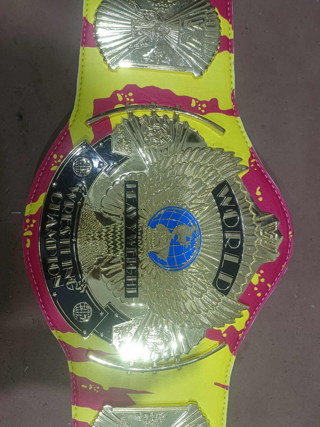 Close-up of a Hulk Hogan championship belt with a gold eagle plate, blue globe and red-yellow strap.
