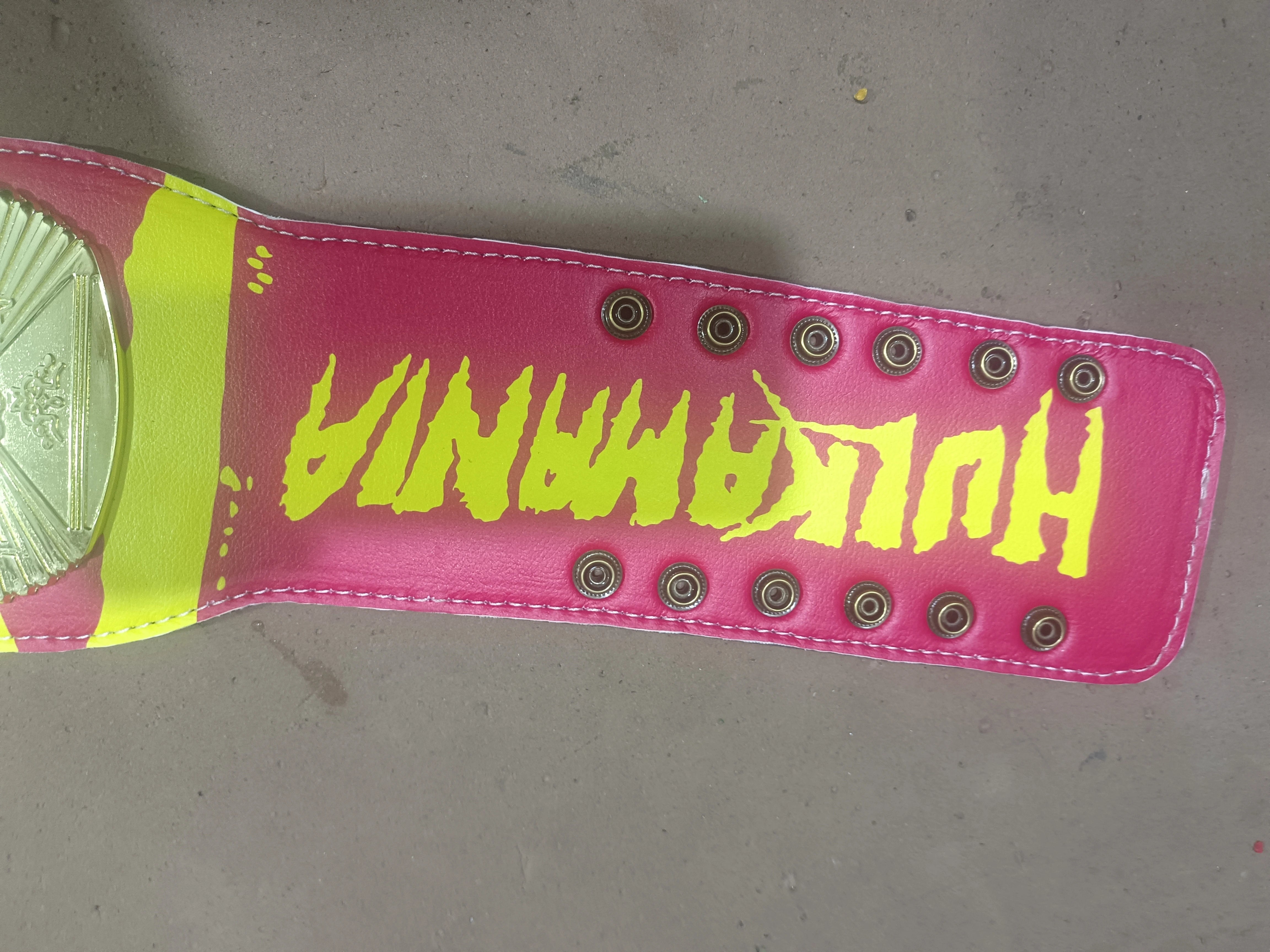 Close-up of a pink Hulkamania belt strap with yellow lettering, metal snaps and white stitched edges.