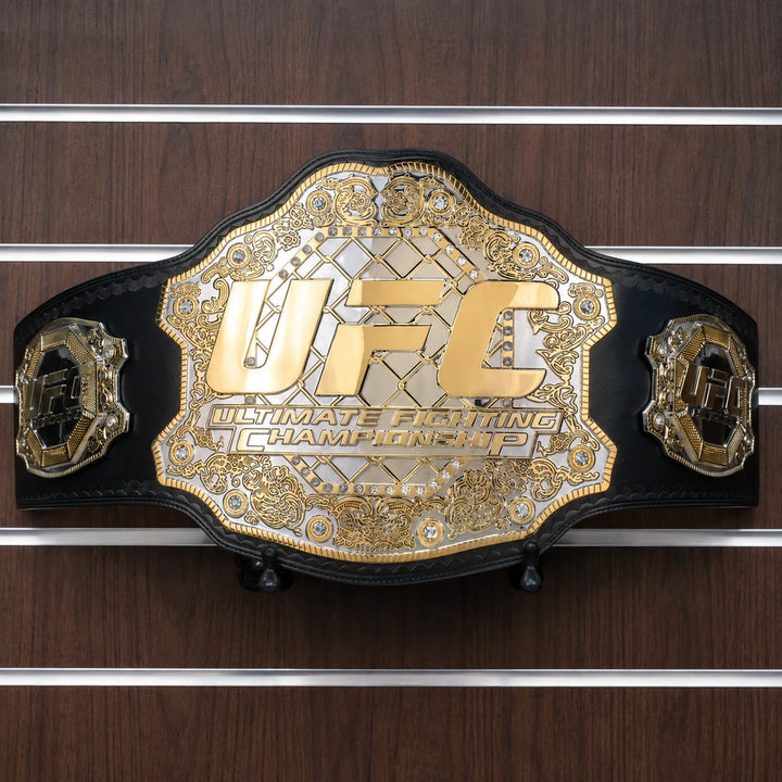 Gold UFC championship belt displayed on a black leather strap against a wooden display wall.